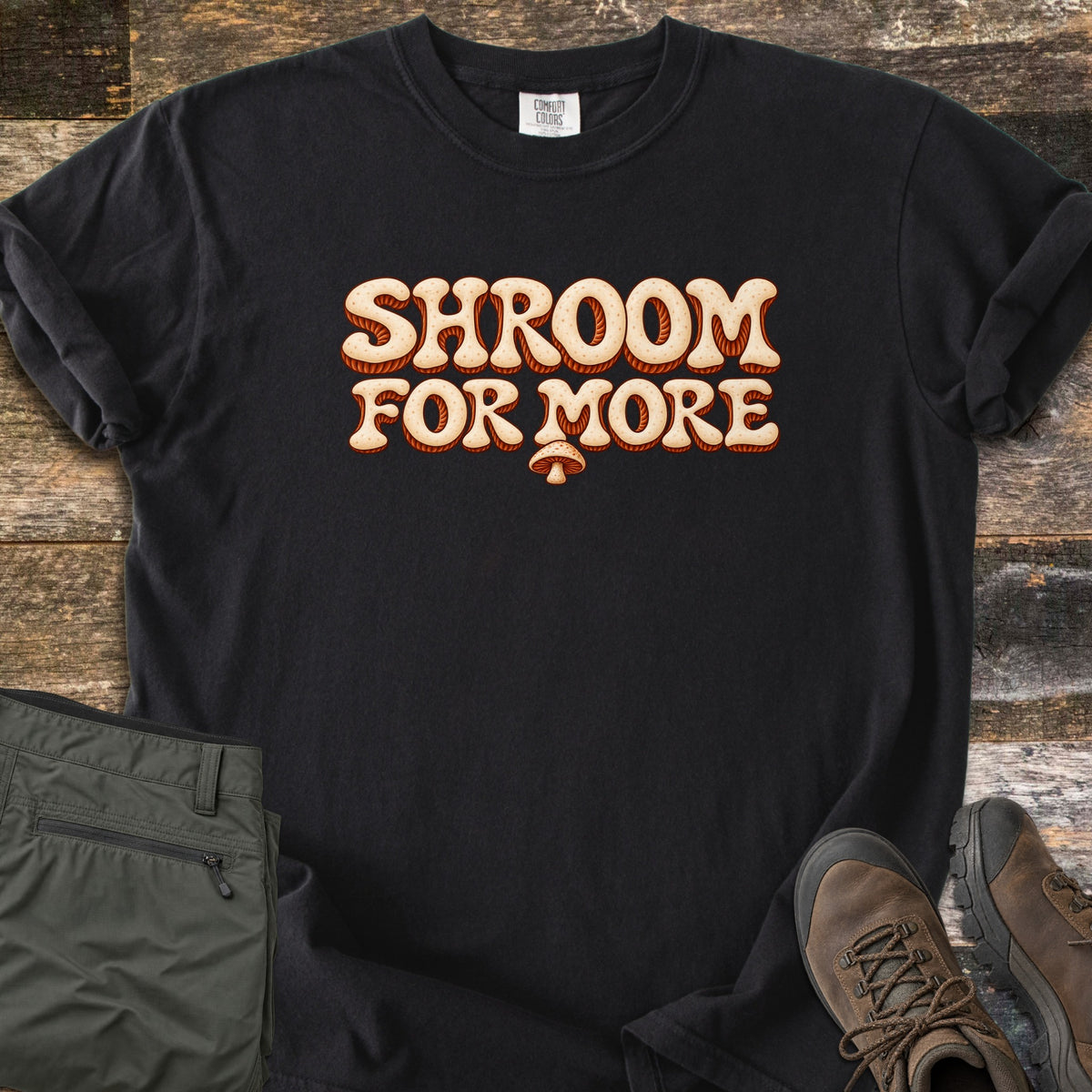 Shroom for More Comfort Colors T-shirt in Black, laid flat. Retro cream bubble letters with rust-orange striped shadows read Shroom For More, with a small mushroom below. Sleeves are rolled on a rustic wood floor beside olive cargo pants and brown hiking boots.