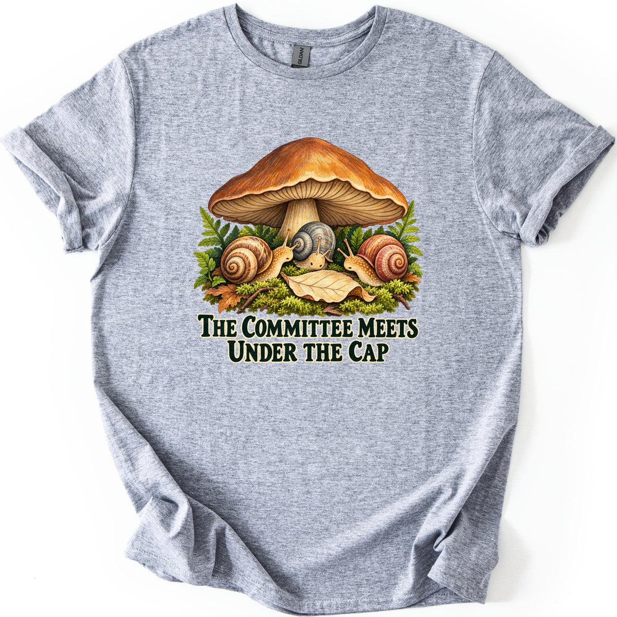 The Committee Meets Under the Cap Comfort Colors T-shirt in Sport Grey, laid flat. A large brown mushroom shelters three snails on moss among ferns and a fallen leaf, above dark green words The Committee Meets Under the Cap. The shirt lies on a white background with sleeves cuffed.