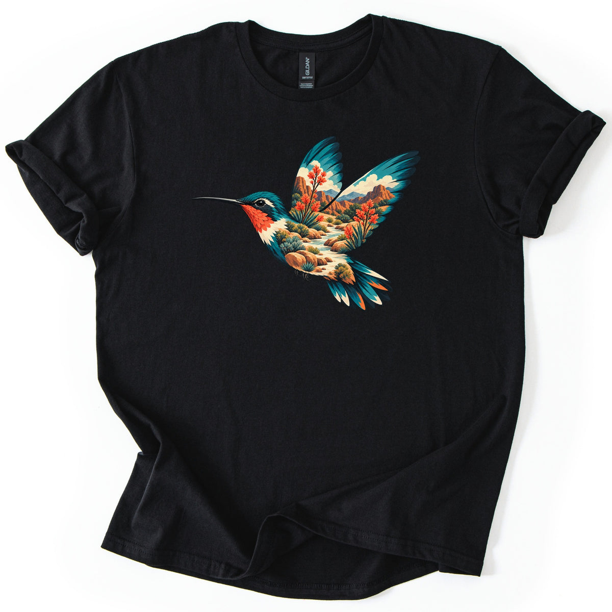 Hummingbird Canyon Comfort Colors T-shirt in Black, laid flat. A hummingbird in flight with teal wings and a red throat, filled with a desert canyon scene of red rocks, clouds, a creek, green shrubs and orange flowering plants. Sleeves cuffed, on a white surface.