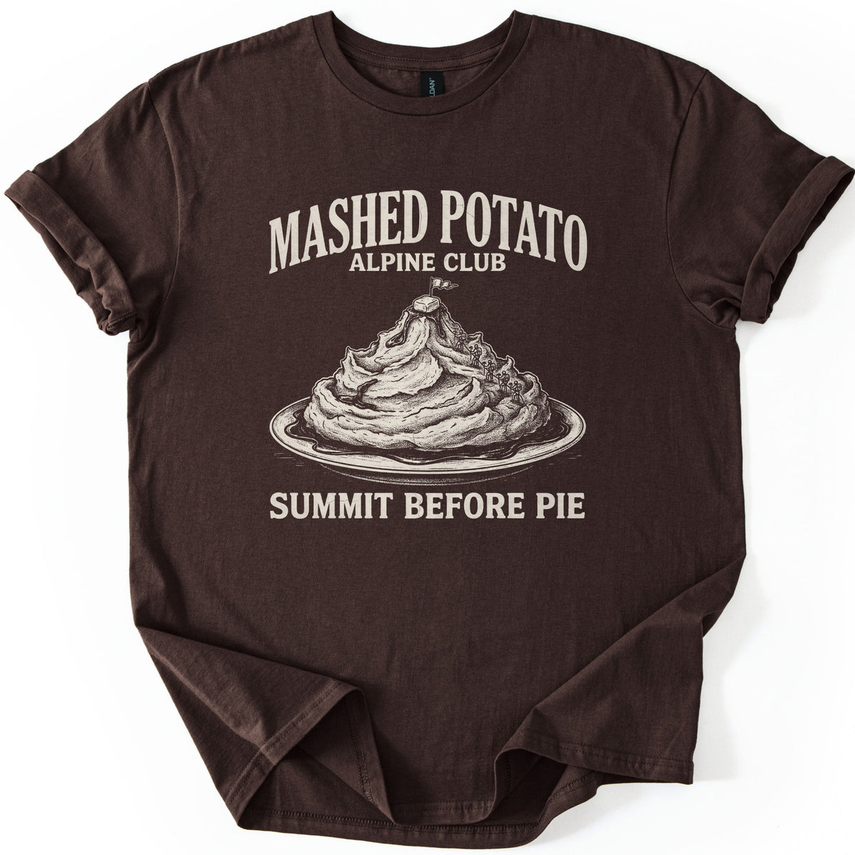 Mashed Potato Alpine Club Comfort Colors T-shirt in Dark Chocolate, laid flat. Cream etched art of a mashed potato mountain on a plate of gravy, with tiny climbers and a butter pat and flag on top, under Mashed Potato Alpine Club and above Summit Before Pie. Rolled sleeves, white background.