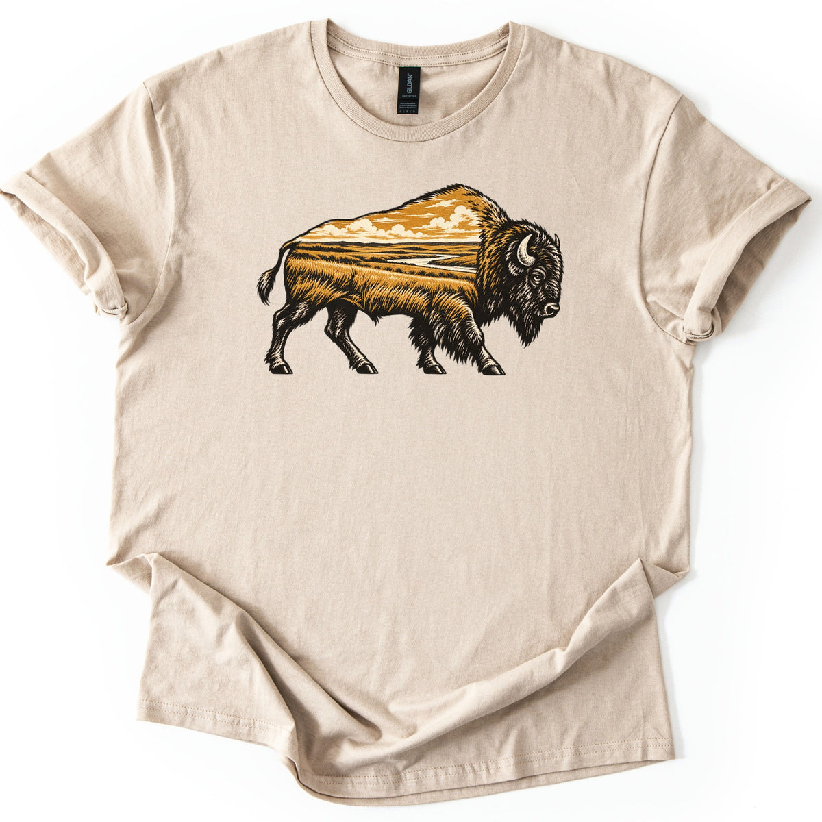 Prairie Bison Comfort Colors T-shirt in Sand, laid flat. A woodcut-style bison walks in side profile, its body filled with a golden prairie scene of tall grass, a winding river and clouds. No words. The shirt has cuffed sleeves on a white background.