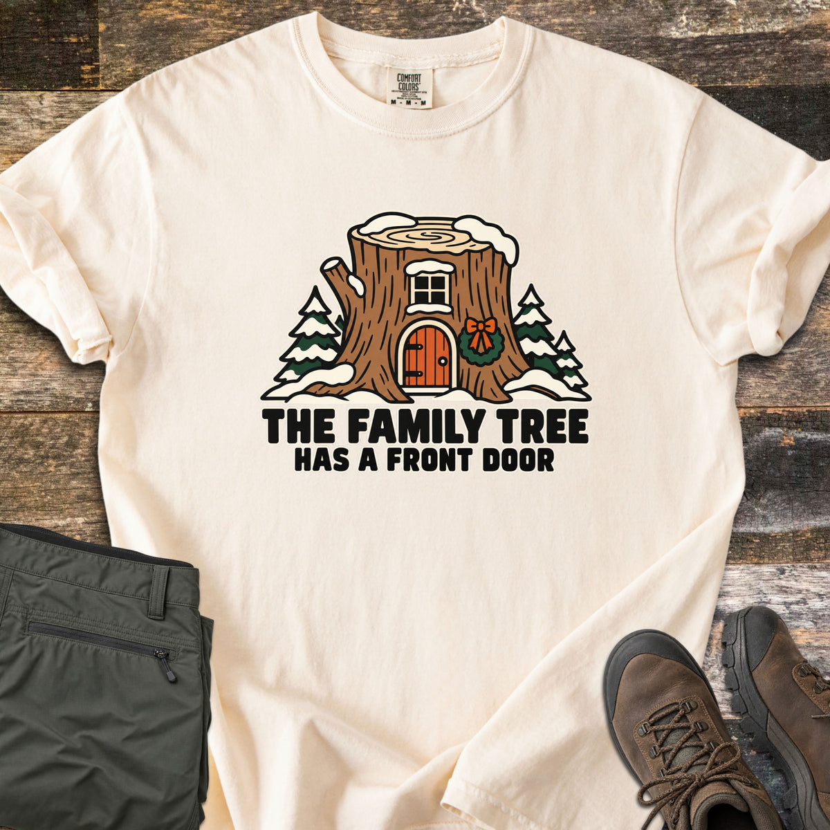 The Family Tree Has a Front Door Comfort Colors T-shirt in Ivory, laid flat. A snowy tree stump house with a window, an orange arched door, and a wreath with a bow sits between pine trees above the words The Family Tree Has a Front Door. Olive pants and brown hiking boots lie on dark wood.