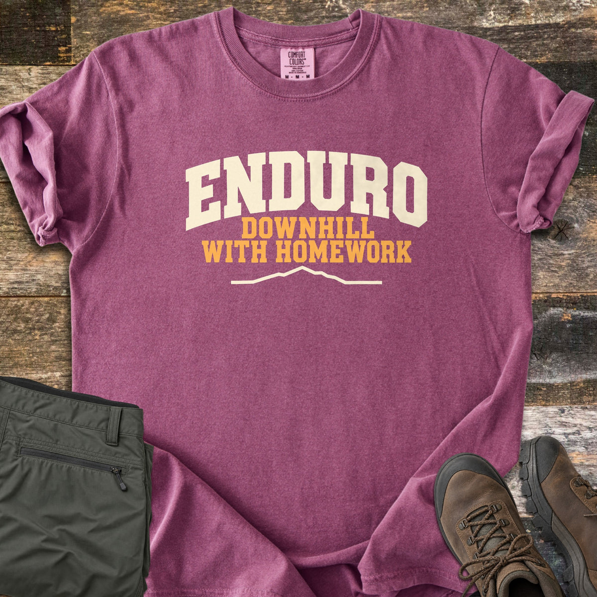 Enduro Downhill With Homework Comfort Colors T-shirt in Berry, laid flat. The word Enduro in large cream arched letters, with Downhill With Homework in orange below and a thin cream mountain ridge line. Sleeves are rolled on a rustic wood surface beside green pants and brown hiking boots.