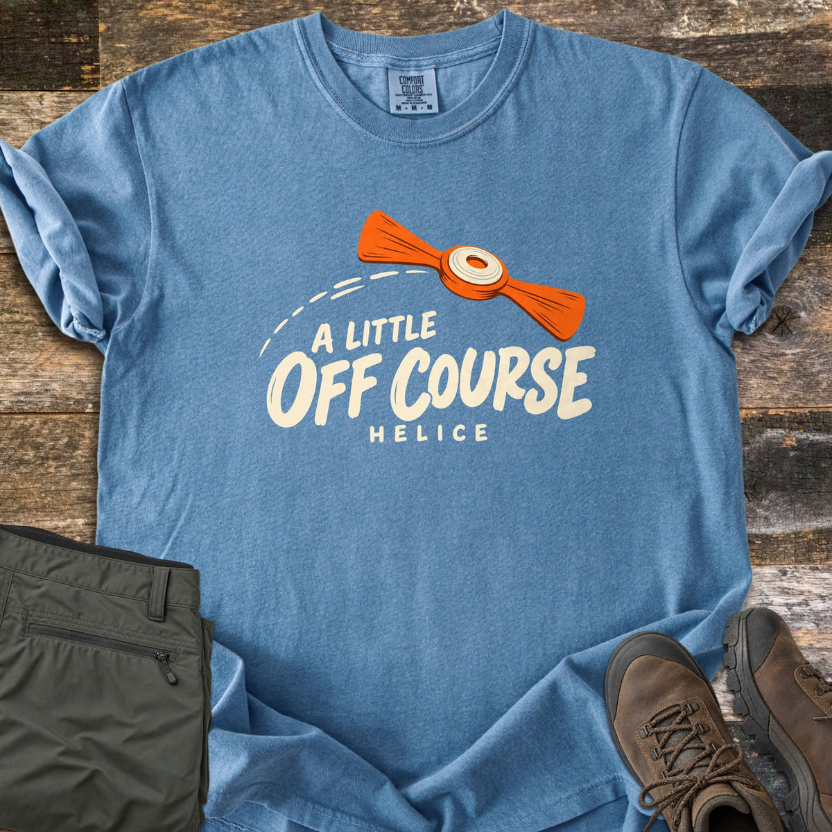 A Little Off Course Comfort Colors T-shirt in Blue Jean, laid flat. An orange helice target with a white center flies along a dashed arc above cream text reading A Little Off Course and Helice. Folded olive hiking pants and brown hiking boots sit beside it on weathered wood.