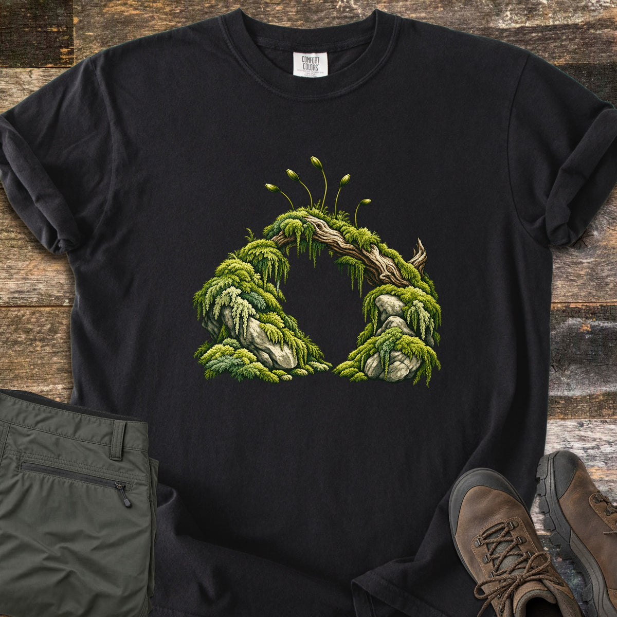 Moss Arch Comfort Colors T-shirt in Black, laid flat. An illustrated arch of moss-covered rocks joined by a weathered branch, with slender moss sporophytes sprouting on top; no words. Sleeves are rolled, on a wood background with olive pants and brown hiking boots.