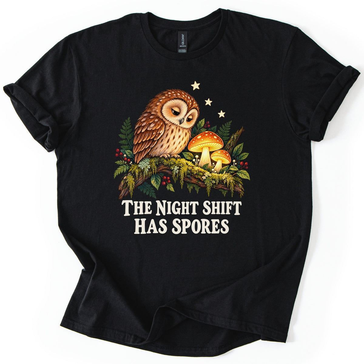 The Night Shift Has Spores Comfort Colors T-shirt in Black, laid flat. A sleepy brown owl perches on a mossy branch beside two glowing orange mushrooms, with ferns, red berries and small stars, above the words The Night Shift Has Spores in cream. Shown on white.
