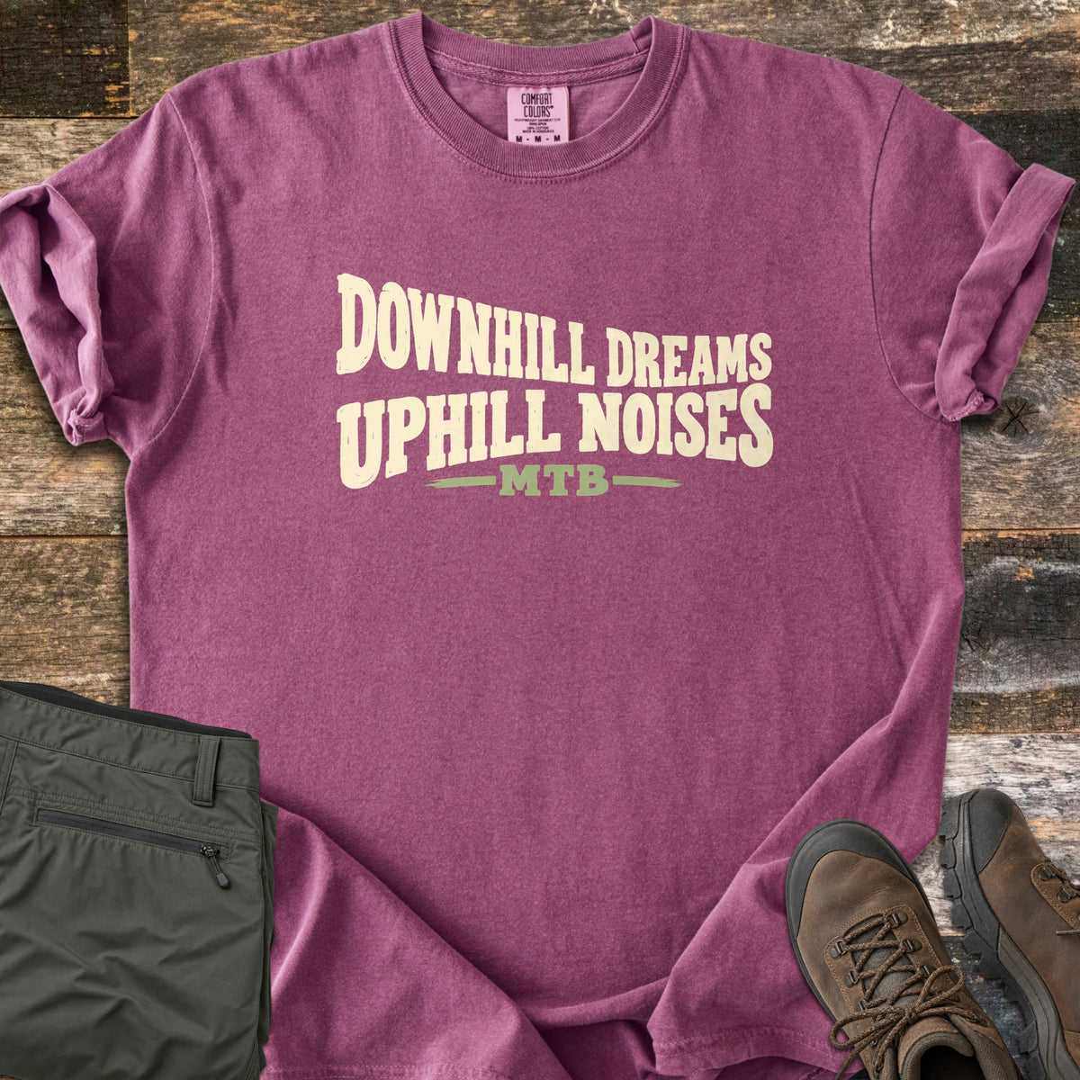 Downhill Dreams Uphill Noises Comfort Colors T-shirt in Berry, laid flat. Cream wavy block letters read Downhill Dreams Uphill Noises with MTB in green between two green lines below. Sleeves rolled on rustic wood planks, with folded olive hiking pants and brown hiking boots.