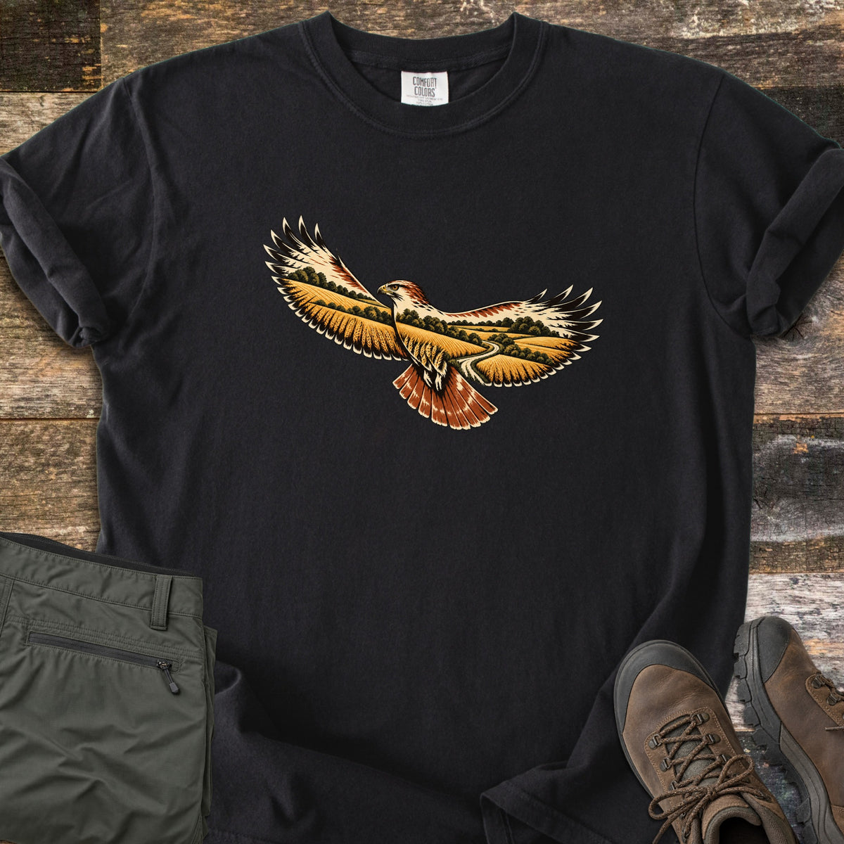 Hawk Over Golden Fields Comfort Colors T-shirt in Black, laid flat. A hawk with outstretched wings and a rust-red tail, its wings filled with golden fields, green trees and a winding path; no words. Folded olive hiking pants and brown hiking boots sit beside it on weathered wood.