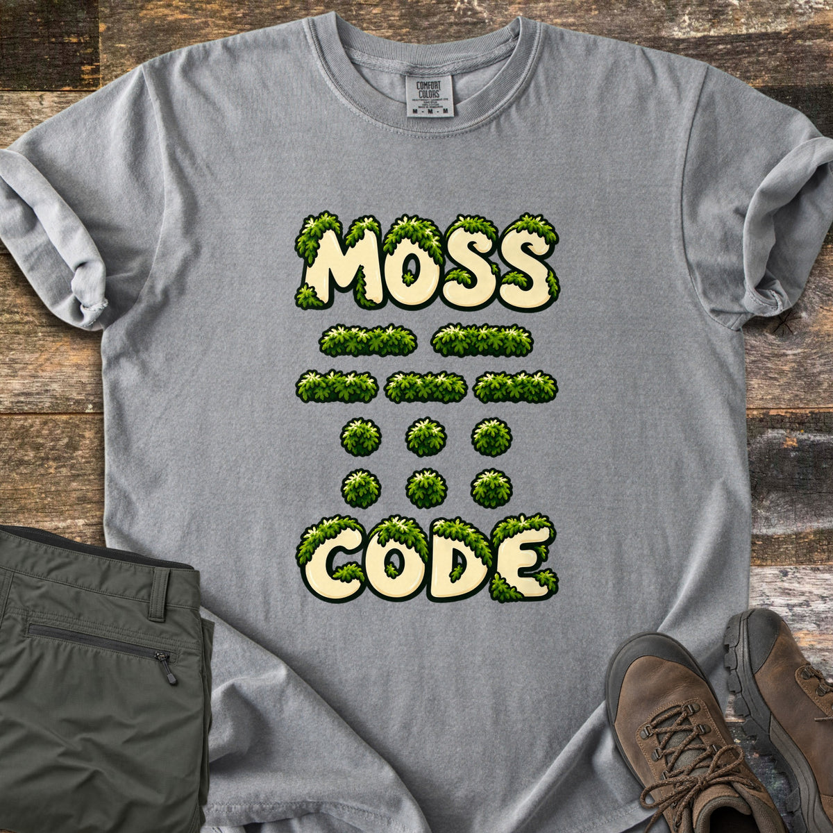 Moss Code Comfort Colors T-shirt in Grey, laid flat. Moss-covered cream bubble letters spell MOSS, then moss-tuft dashes and dots spelling MOSS in Morse code, then CODE. It lies on weathered wood beside folded olive hiking pants and a brown hiking boot.