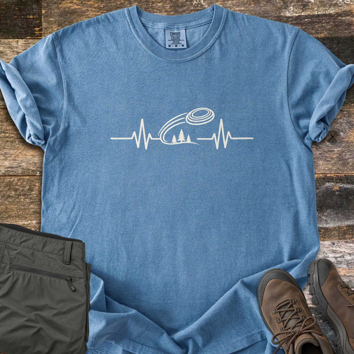 Trap Shooting Heartbeat Comfort Colors T-shirt in Blue Jean, laid flat. A cream heartbeat line rises into a flying clay target with motion lines above three pine trees, with no words. Sleeves are rolled on weathered wood beside folded olive pants and brown hiking boots.