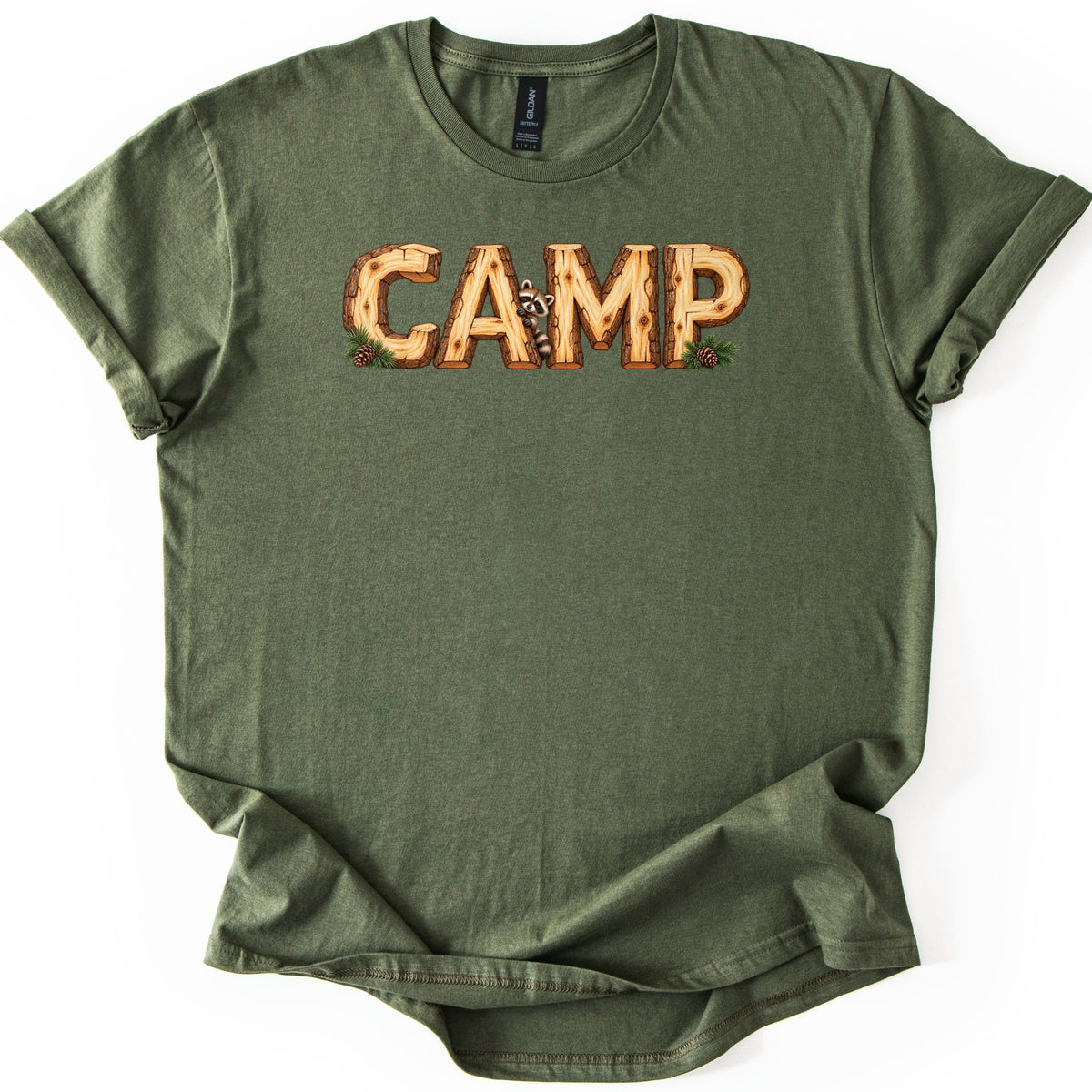Camp Comfort Colors T-shirt in Military Green, laid flat. The word CAMP is printed in letters shaped from wooden logs with bark trim, a raccoon peeking between the A and M, and pinecones on pine sprigs at each end. The shirt lies on white with rolled sleeves.
