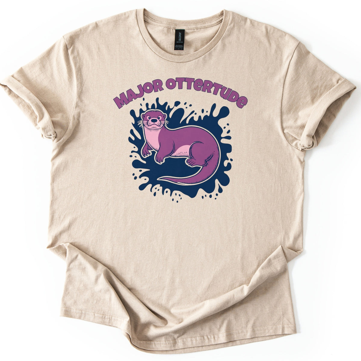 Major Ottertude Comfort Colors T-shirt in Sand, laid flat. A purple cartoon otter with a pink chest rests on a navy blue paint splash beneath arched purple letters reading MAJOR OTTERTUDE. The shirt has cuffed sleeves and lies on a white background.