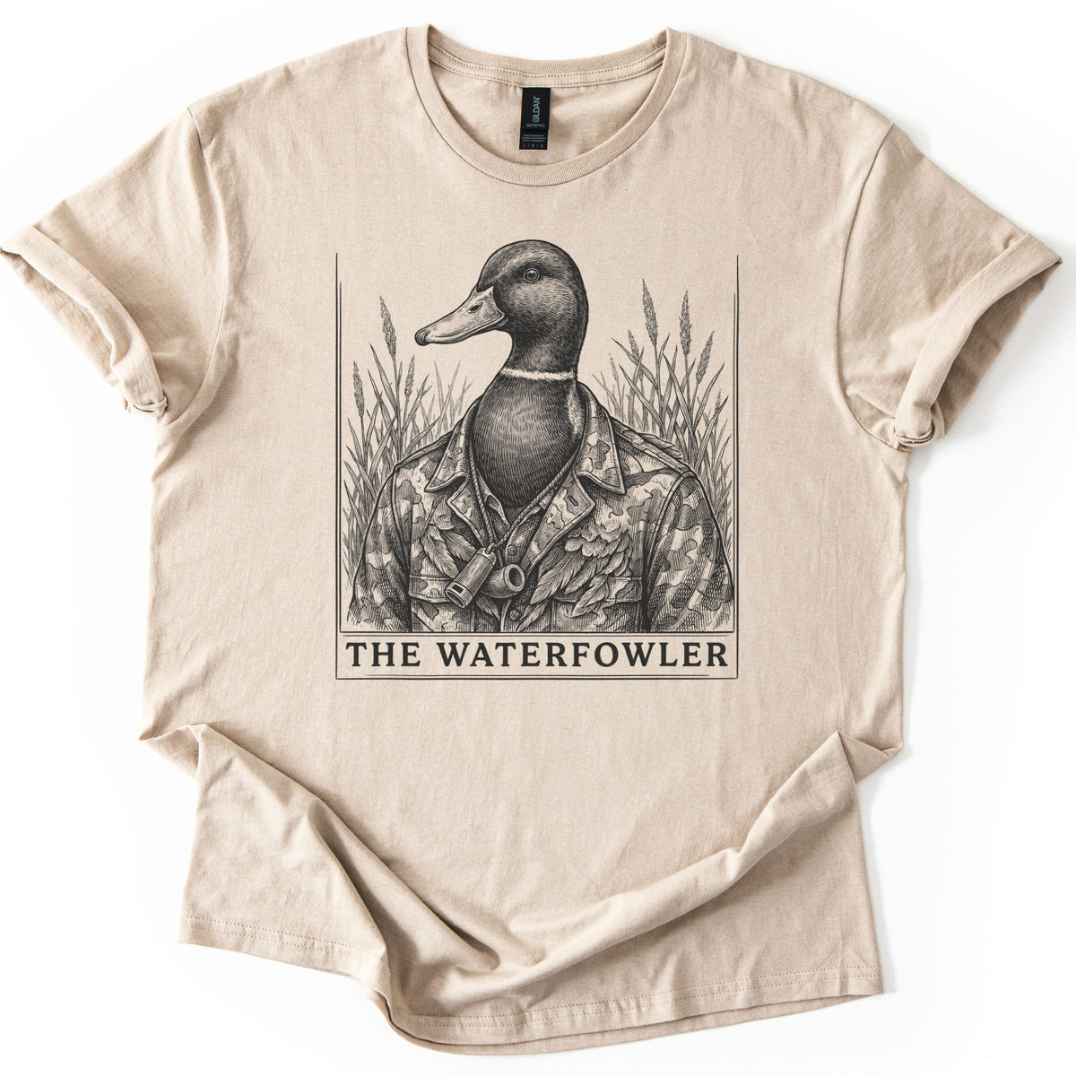 The Waterfowler Comfort Colors T-shirt in Sand, laid flat. A black engraving-style drawing of a mallard duck in a camo shirt with a duck call on a lanyard, set in tall marsh grass inside a thin frame, above the words THE WATERFOWLER. Sleeves cuffed, on white.