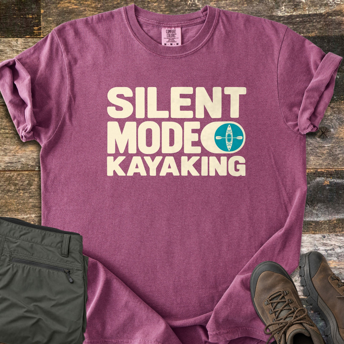 Silent Mode Comfort Colors T-shirt in Berry, laid flat. Cream block letters read SILENT MODE KAYAKING, with a toggle switch icon showing a teal circle with a kayak and paddle. Sleeves are rolled on a wood background beside folded olive pants and brown hiking boots.