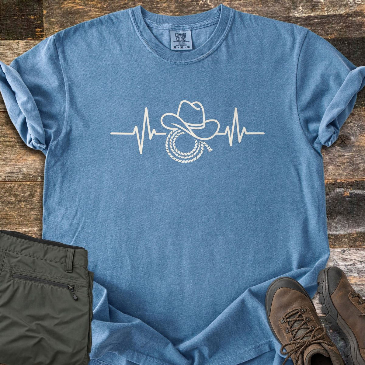 Cowboy Heartbeat Comfort Colors T-shirt in Blue Jean, laid flat. A cream line art heartbeat line rises into peaks around a cowboy hat resting on a coiled lasso; there are no words. Sleeves are cuffed, on rustic wood boards beside folded olive cargo pants and brown hiking boots.