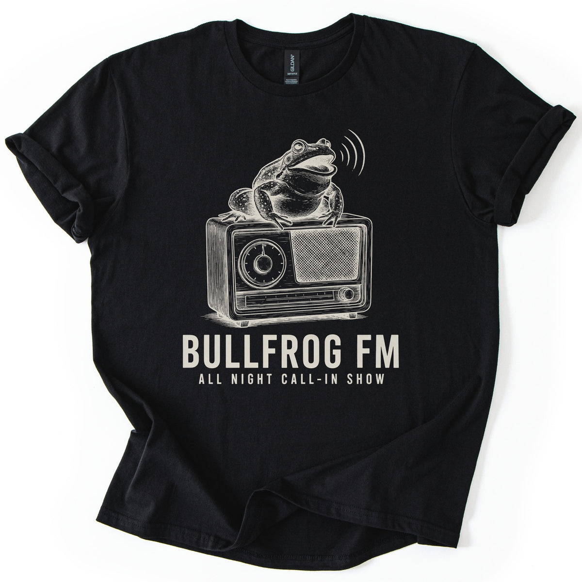 Bullfrog FM Comfort Colors T-shirt in Black, laid flat. A cream ink sketch of a croaking bullfrog on a vintage radio, sound waves at its mouth, above the words BULLFROG FM and ALL NIGHT CALL-IN SHOW. Sleeves are cuffed on a white background.