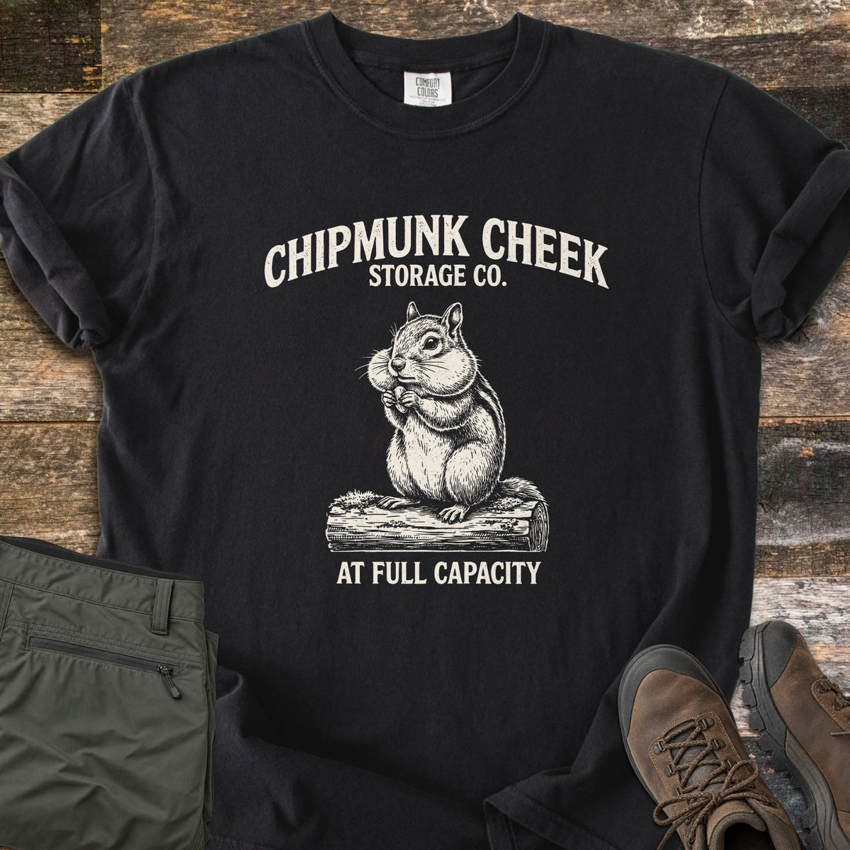 Chipmunk Cheek Storage Co Comfort Colors T-shirt in Black, laid flat. Cream print of a chipmunk with full cheeks sitting on a log, under the words Chipmunk Cheek Storage Co. and above At Full Capacity. Rolled sleeves, on weathered wood with olive pants and brown hiking boots.