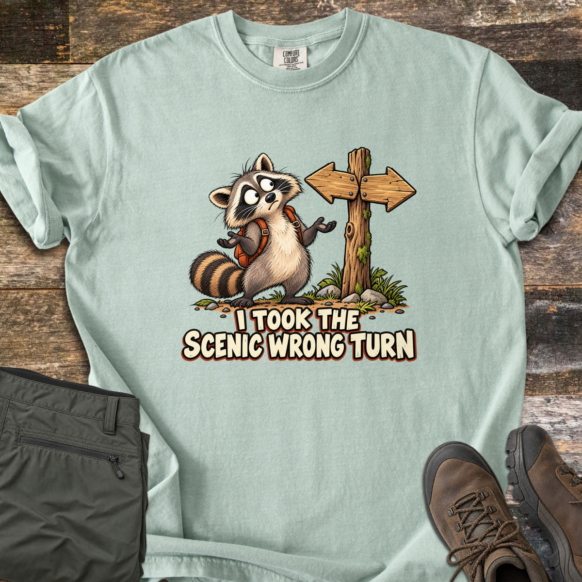 I Took the Scenic Wrong Turn Comfort Colors T-shirt in Bay, laid flat. A cartoon raccoon with a backpack shrugs, looking confused beside a wooden signpost with arrows pointing both ways, above the words I Took the Scenic Wrong Turn. Olive hiking pants and brown boots lie on weathered wood.