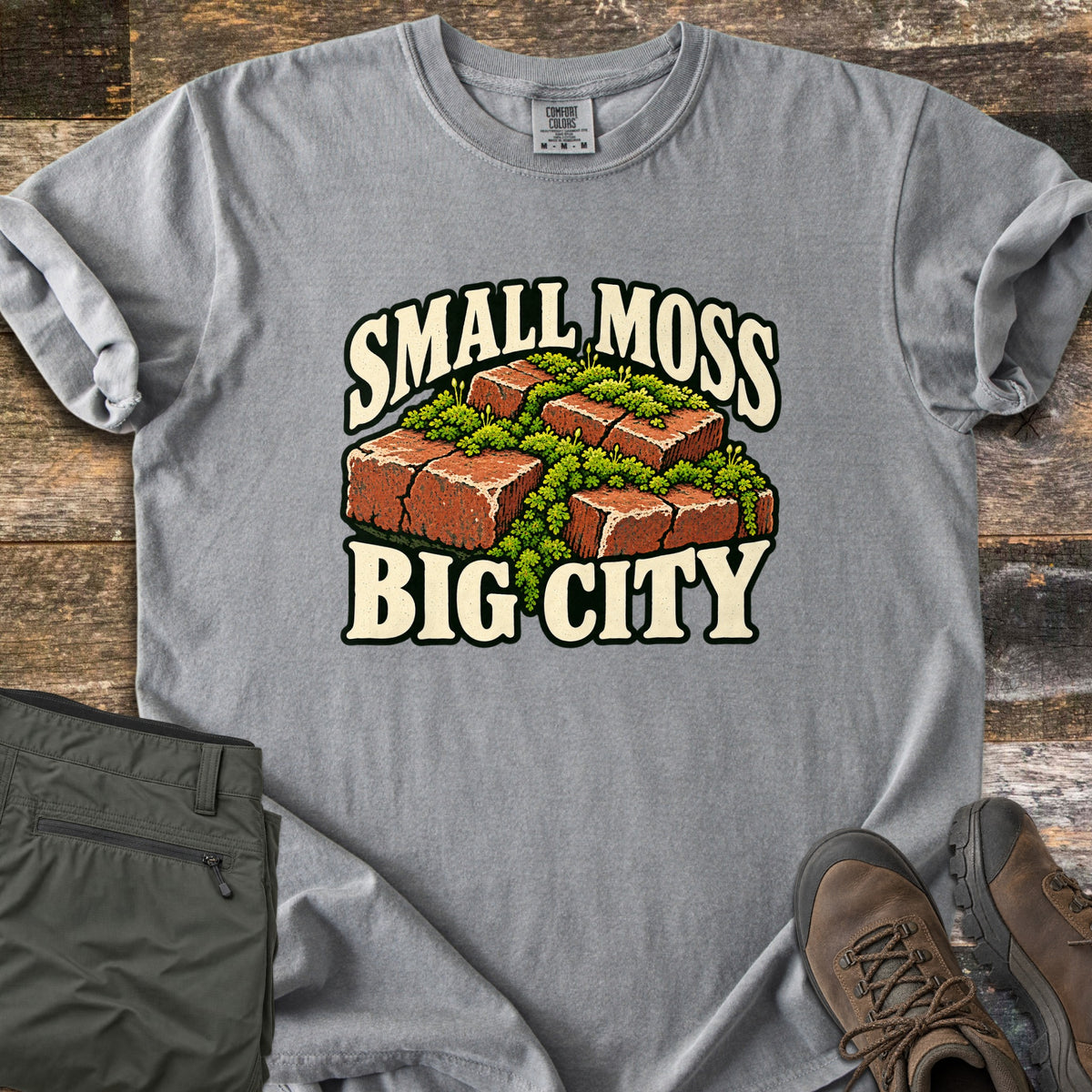 Small Moss Big City Comfort Colors T-shirt in Grey, laid flat. Red bricks with bright green moss growing between and over them, with the words Small Moss Big City in cream lettering. Sleeves are rolled, on a wood floor beside folded olive pants and brown hiking boots.