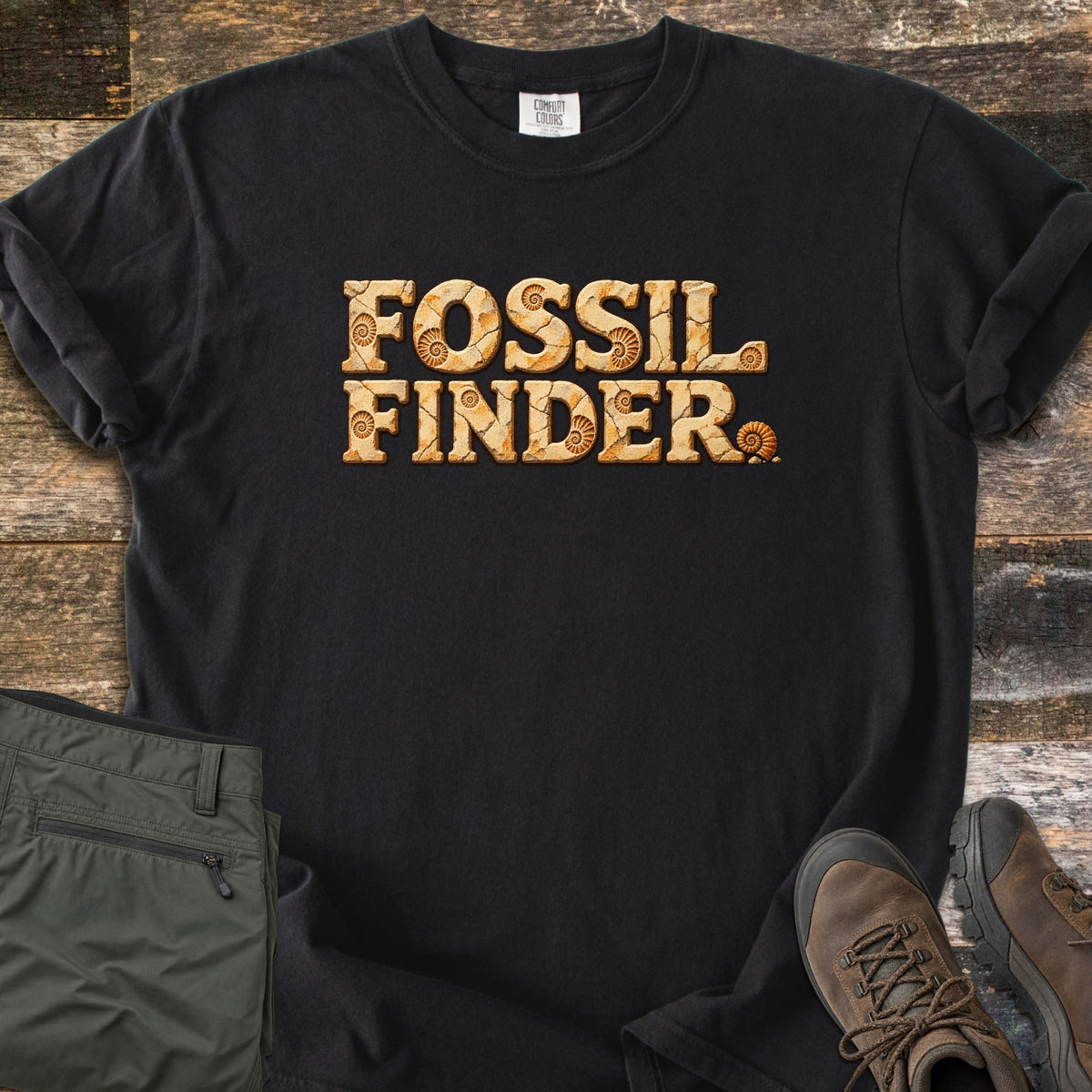 Fossil Finder Comfort Colors T-shirt in Black, laid flat. The words Fossil Finder are printed in cracked tan stone letters with ammonite shell fossils, plus a small ammonite after the last letter. Sleeves are rolled, on a rustic wood floor with olive cargo pants and brown hiking boots.