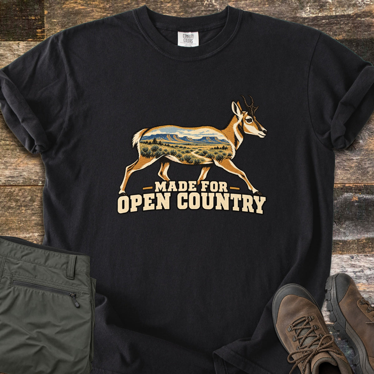 Made for Open Country Comfort Colors T-shirt in Black, laid flat. A running pronghorn whose body is filled with a desert scene of mesas, sagebrush and a trail, above the words Made for Open Country. Sleeves are rolled on a wood background with green pants and brown hiking boots.