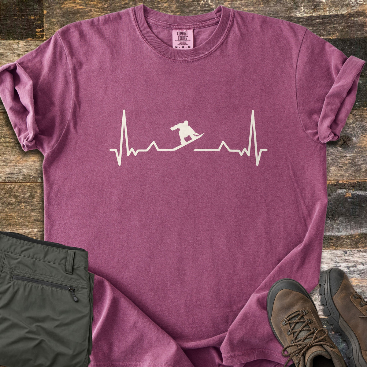 Snowboarder Heartbeat Comfort Colors T-shirt in Berry, laid flat. A cream heartbeat line runs across the chest, with a snowboarder silhouette riding the line in the middle. Sleeves are rolled, on a rustic wood floor beside folded olive pants and brown hiking boots.