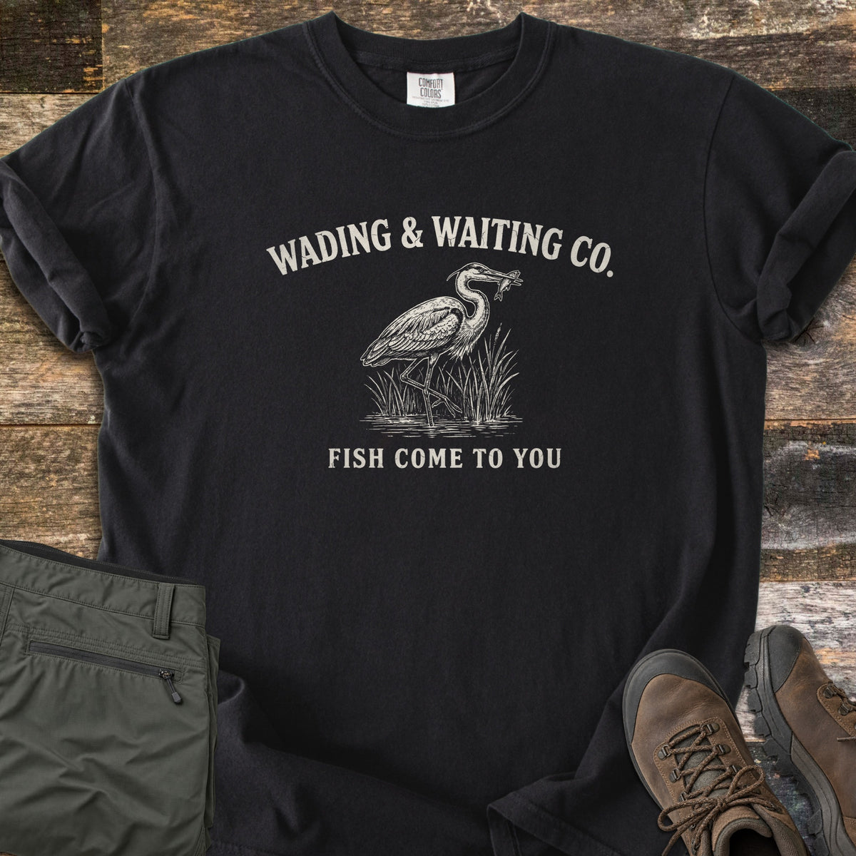 Wading & Waiting Co. Comfort Colors T-shirt in Black, laid flat. A heron stands in marsh reeds holding a fish in its beak, under the arched words Wading & Waiting Co. and above Fish Come To You. Olive cargo pants and brown hiking boots lie beside it on weathered wood.