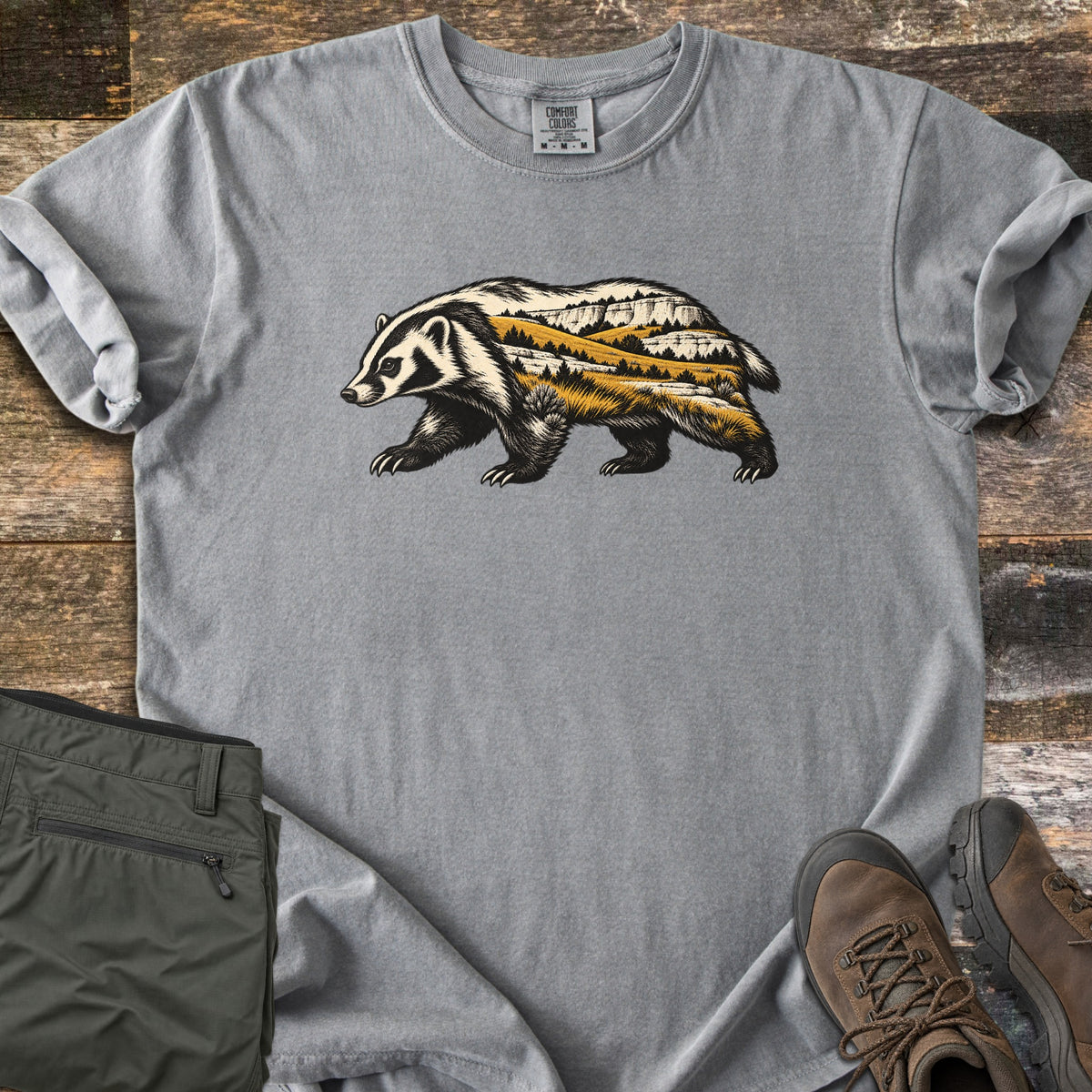 Badger Prairie Bluffs Comfort Colors T-shirt in Grey, laid flat. A walking badger silhouette filled with a prairie scene of golden grass, pine trees and white bluffs is printed on the chest. Shown on weathered wood with folded olive pants and brown hiking boots.