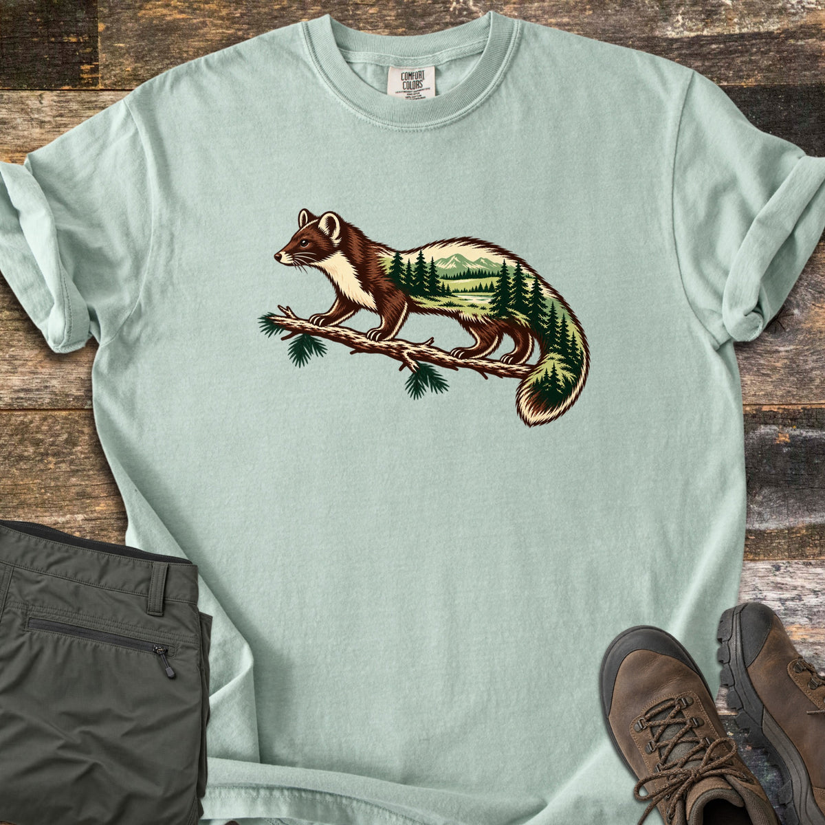 Pine Marten Forest Comfort Colors T-shirt in Bay, laid flat. A pine marten stands on a pine branch, its body filled with a scene of evergreen trees, a lake and mountains. No words are printed. Shown on weathered wood with folded olive pants and brown hiking boots.