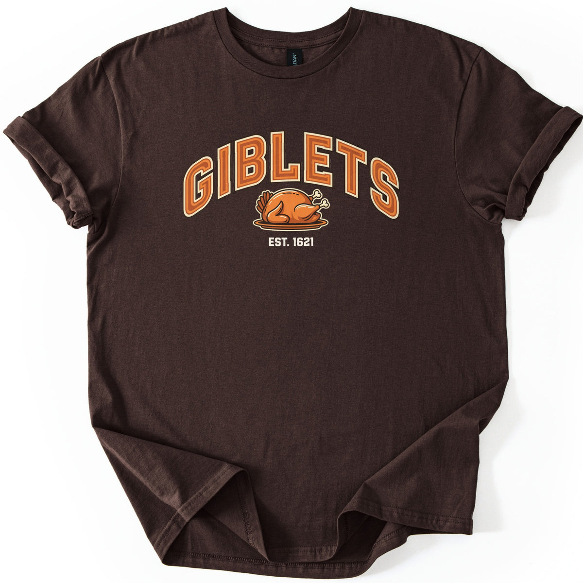 Giblets Comfort Colors T-shirt in Dark Chocolate, laid flat. Arched orange varsity-style letters read GIBLETS above a cartoon roast turkey on a platter, with EST. 1621 in white below. The shirt has rolled sleeves and lies on a white background.