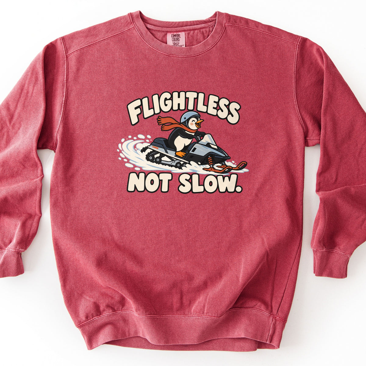 Flightless Not Slow Comfort Colors crewneck sweatshirt in Crimson, laid flat. A smiling cartoon penguin in a helmet and red scarf rides a snowmobile kicking up snow, with Flightless printed above and Not Slow. below in cream letters. It lies on a white background.