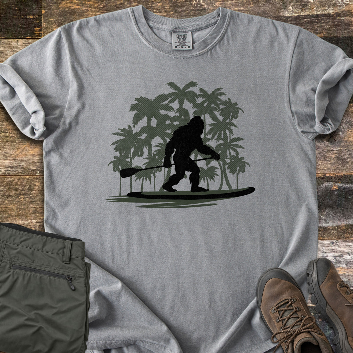 Paddle Board Squatch Comfort Colors T-shirt in Grey, laid flat. A black silhouette of Bigfoot standing on a paddle board and holding a paddle, in front of dark green palm trees. Olive pants and brown hiking boots lie beside the shirt on weathered wood planks.
