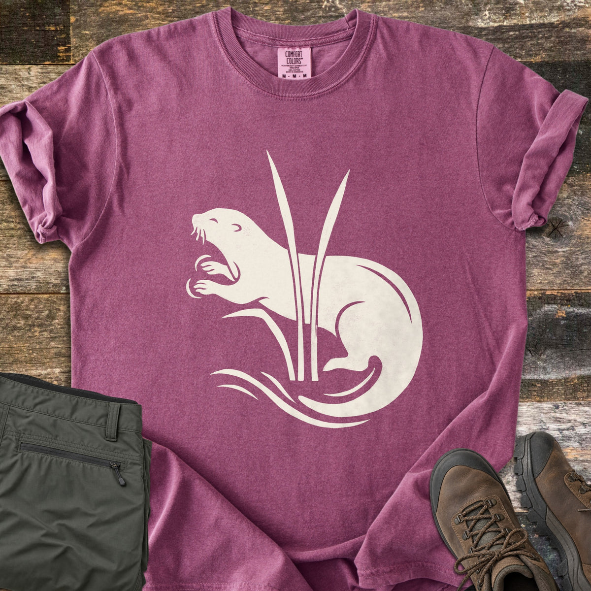 Otter In The Reeds Comfort Colors T-shirt in Berry, laid flat. A cream silhouette of an otter with open mouth and raised paws curls behind two tall reeds above wavy water lines, no words. Folded olive hiking pants and brown hiking boots lie on weathered wood.