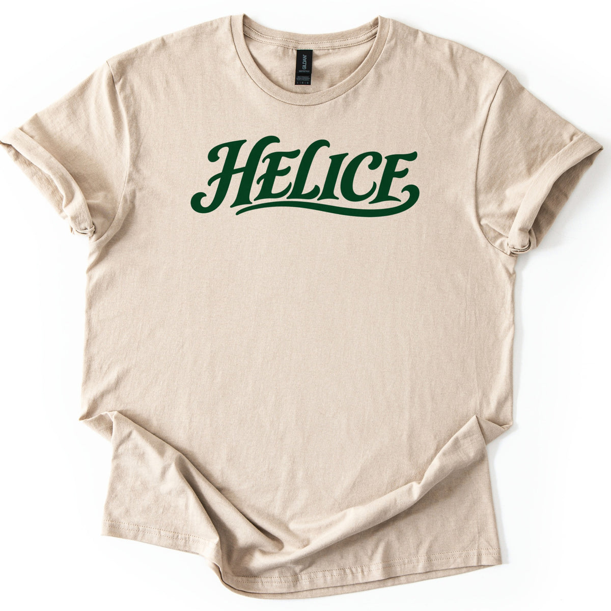 Helice Comfort Colors T-shirt in Sand, laid flat. The word Helice is printed across the chest in dark green retro script with a swooping underline tail. The shirt lies on a white background with its sleeves cuffed and the hem loosely folded.