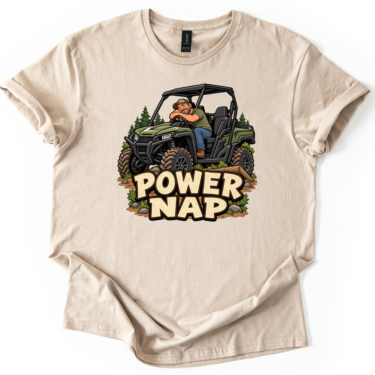 Power Nap Comfort Colors T-shirt in Sand, laid flat. A cartoon man sleeps slumped over the wheel of a green side-by-side UTV parked by a log and pine trees, above the words Power Nap in cream letters. The shirt lies on a white background with sleeves cuffed.