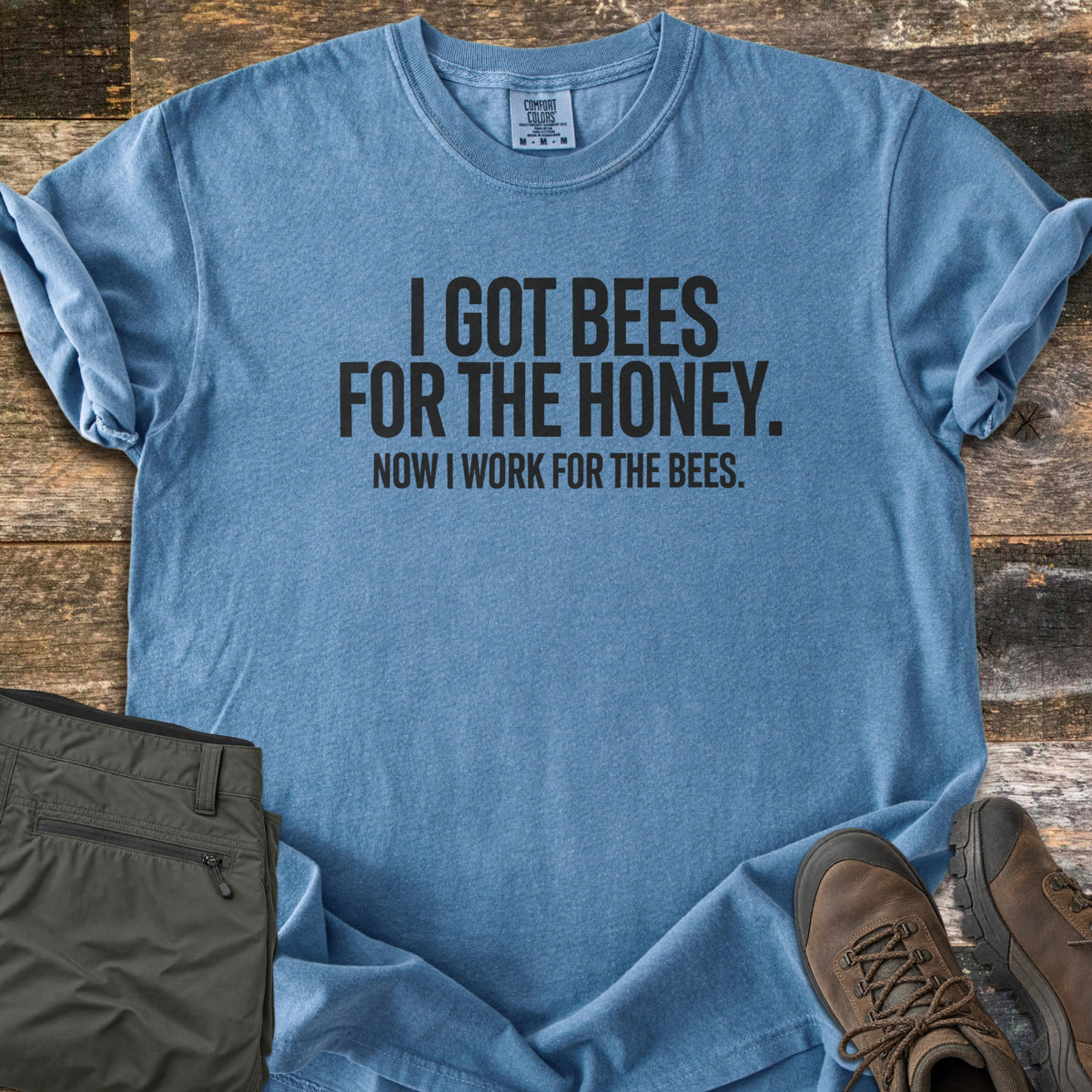 Work For The Bees Comfort Colors T-shirt in Blue Jean, laid flat. Black block letters read I GOT BEES FOR THE HONEY. NOW I WORK FOR THE BEES. The shirt lies with cuffed sleeves on weathered wood planks beside folded olive green pants and brown hiking boots.