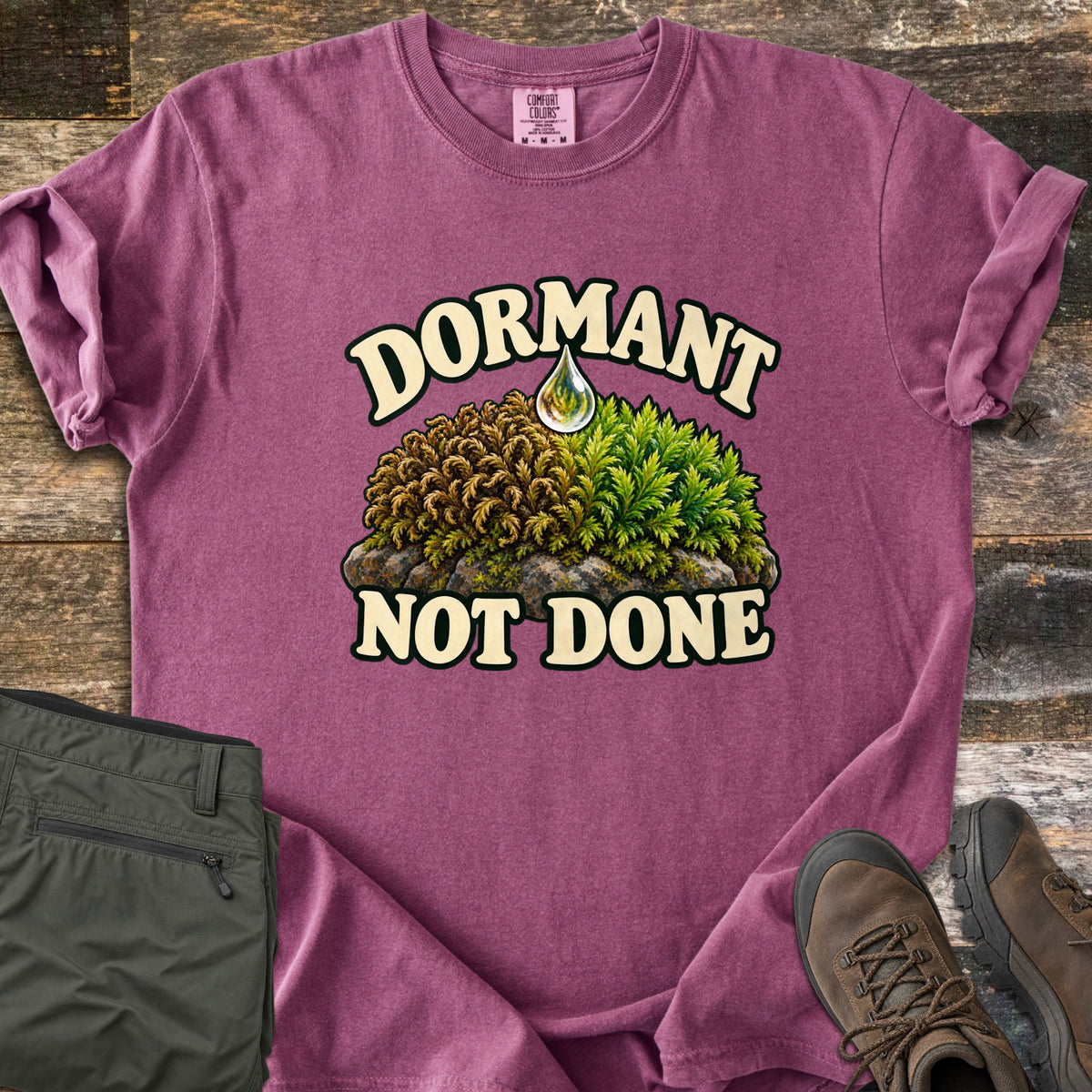 Dormant Not Done Comfort Colors T-shirt in Berry, laid flat. Cream letters spell Dormant above a clump of moss on a rock that turns from dry brown to green, with a water drop at the top and Not Done beneath. Olive pants and brown hiking boots rest on old wood boards.