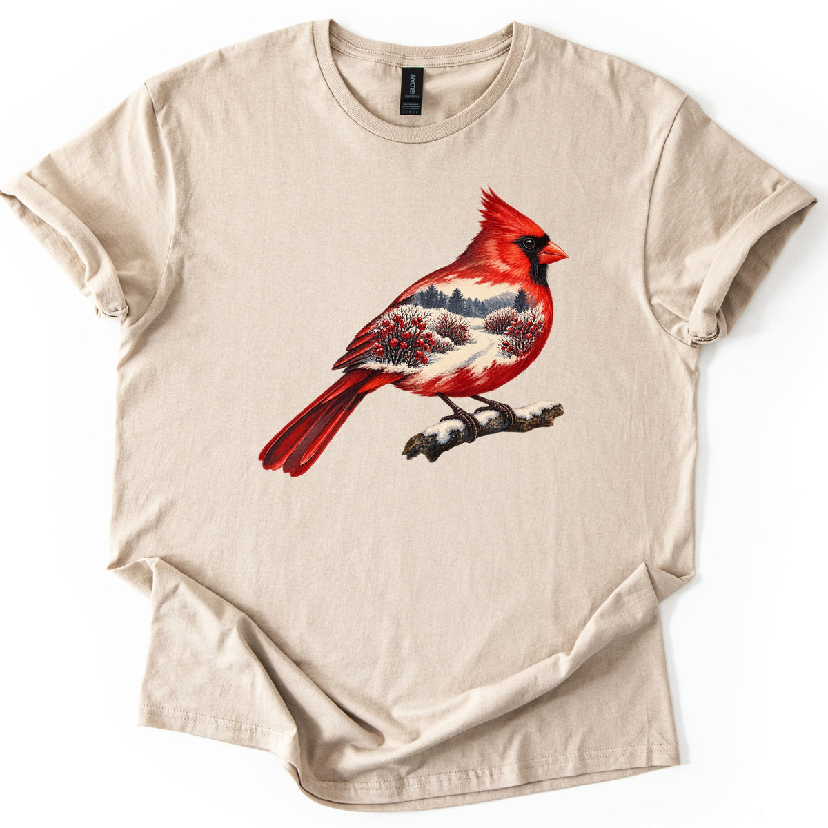Winter Cardinal Comfort Colors T-shirt in Sand, laid flat. A painted red cardinal perches on a snowy branch, its body filled with a winter scene of evergreens, a snowy path and red-berry bushes. No words are printed. The sleeves are cuffed on a white background.