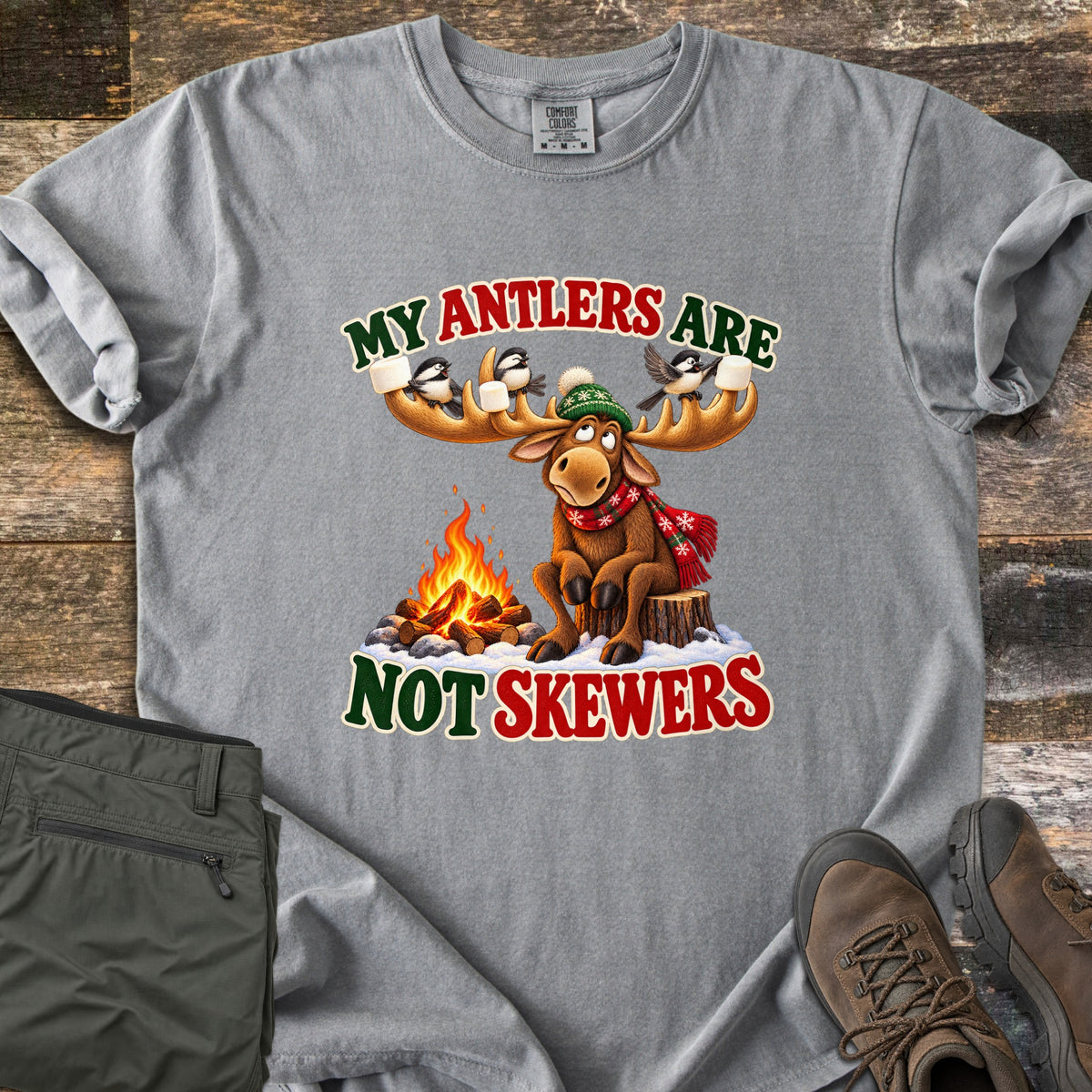 My Antlers Are Not Skewers Comfort Colors T-shirt in Grey, laid flat. A cartoon moose in a green knit hat and red scarf sits on a stump by a campfire as chickadees on its antlers hold marshmallows, with the words My Antlers Are Not Skewers. Olive pants and brown boots lie on weathered wood.