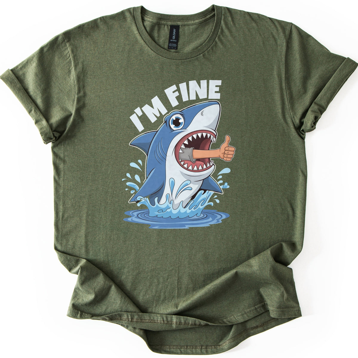 Shark I'm Fine Comfort Colors T-shirt in Military Green, laid flat. A cartoon blue shark leaps from splashing water with a human arm giving a thumbs up sticking out of its open mouth, under the words I'M FINE in white. The shirt has rolled sleeves on a white background.