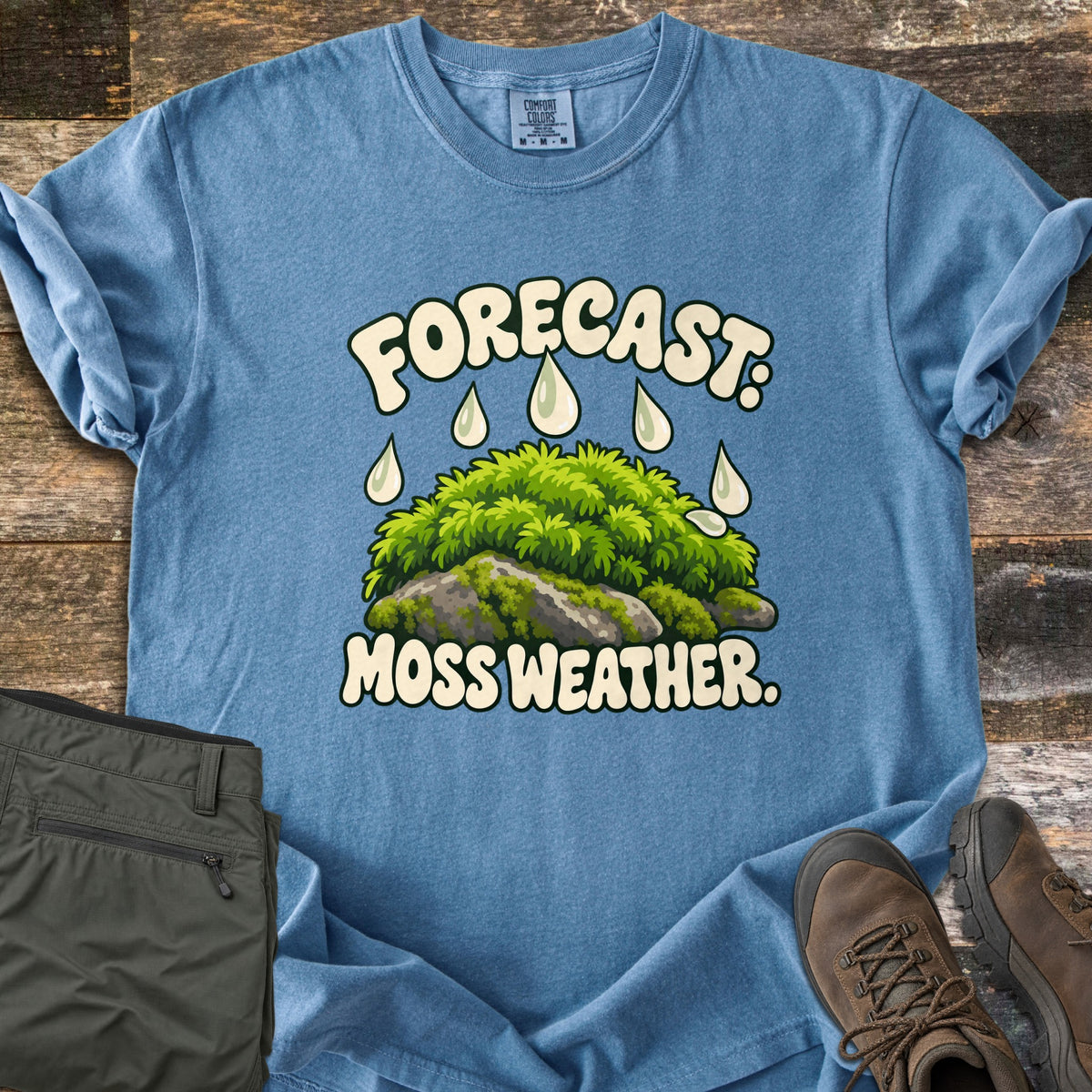 Forecast Moss Weather Comfort Colors T-shirt in Blue Jean, laid flat. A cartoon mound of bright green moss on a gray rock, with raindrops falling from the words Forecast: above and Moss Weather. below. Olive hiking pants and brown hiking boots sit beside it on weathered wood boards.