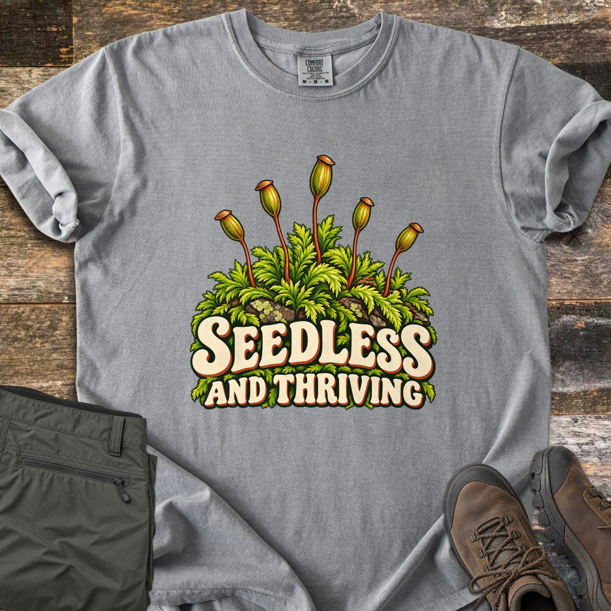 Seedless and Thriving Comfort Colors T-shirt in Grey, laid flat. A clump of green moss with slender red stalks capped by spore capsules rises above the words Seedless and Thriving in cream retro letters. Folded olive pants and brown hiking boots sit beside it on weathered wood.