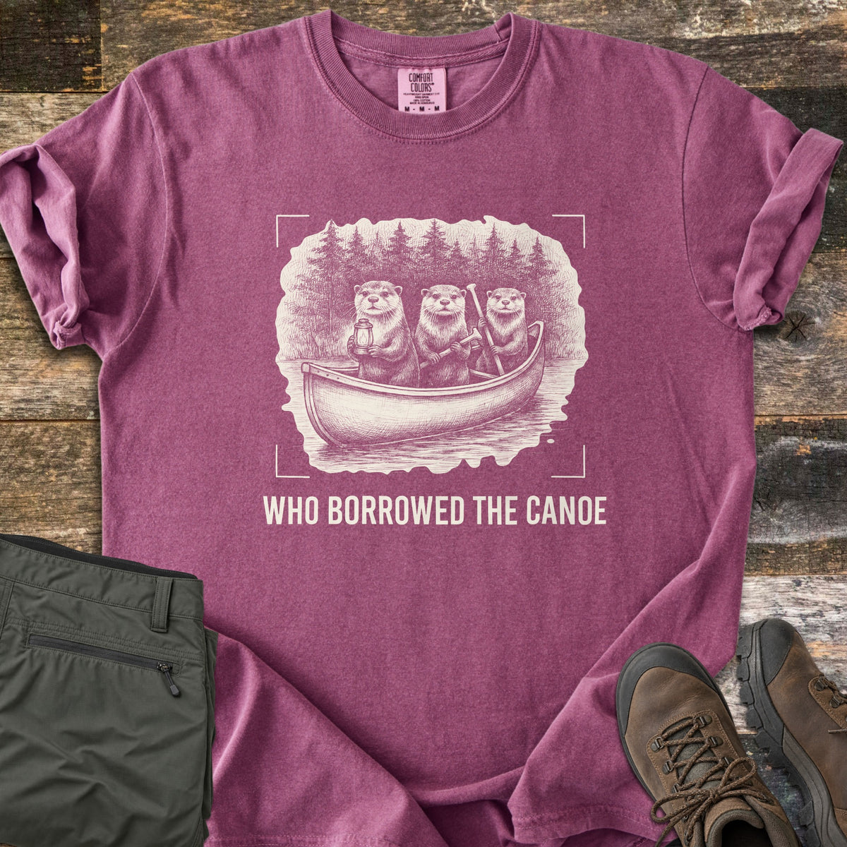 Who Borrowed The Canoe Comfort Colors T-shirt in Berry, laid flat. A cream sketch of three otters paddling a canoe before pine trees, one holding a lantern, inside camera corner marks above the words Who Borrowed The Canoe. Olive hiking pants and brown boots lie on wood.