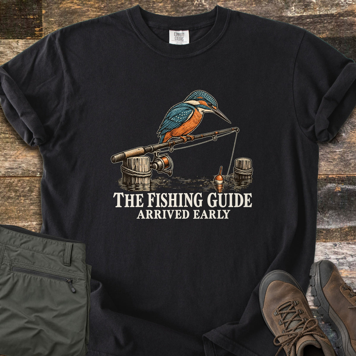 The Fishing Guide Arrived Early Comfort Colors T-shirt in Black, laid flat. A kingfisher perches on a fishing rod resting on rope-wrapped dock posts, its line and orange bobber in the water, above the words The Fishing Guide Arrived Early in cream. Olive pants and brown hiking boots lie on weathered wood.