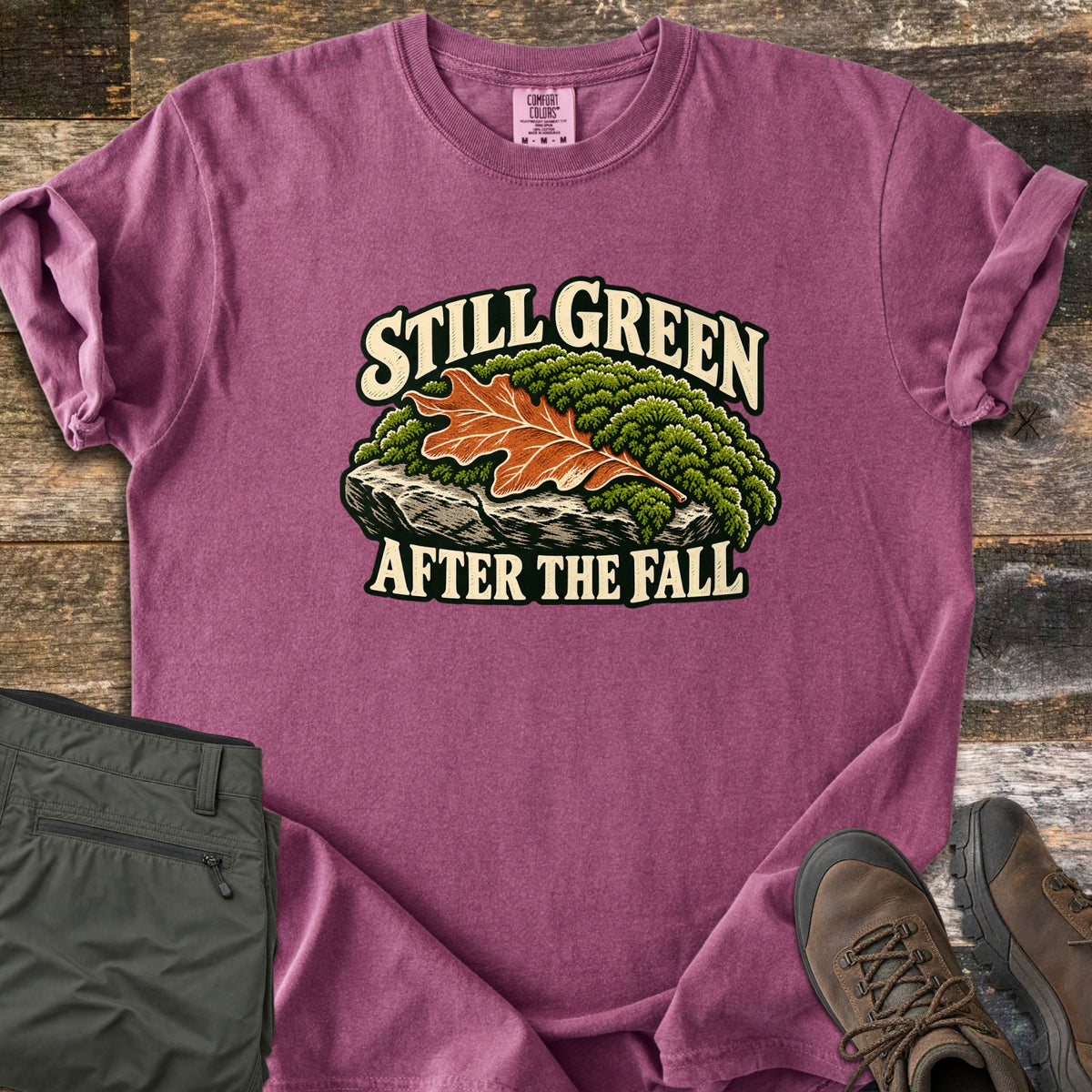Still Green After the Fall Comfort Colors T-shirt in Berry, laid flat. An orange oak leaf rests on a mound of green moss on a gray rock, framed by the words Still Green above and After the Fall below in cream letters. Olive hiking pants and brown boots lie beside it on old wood.