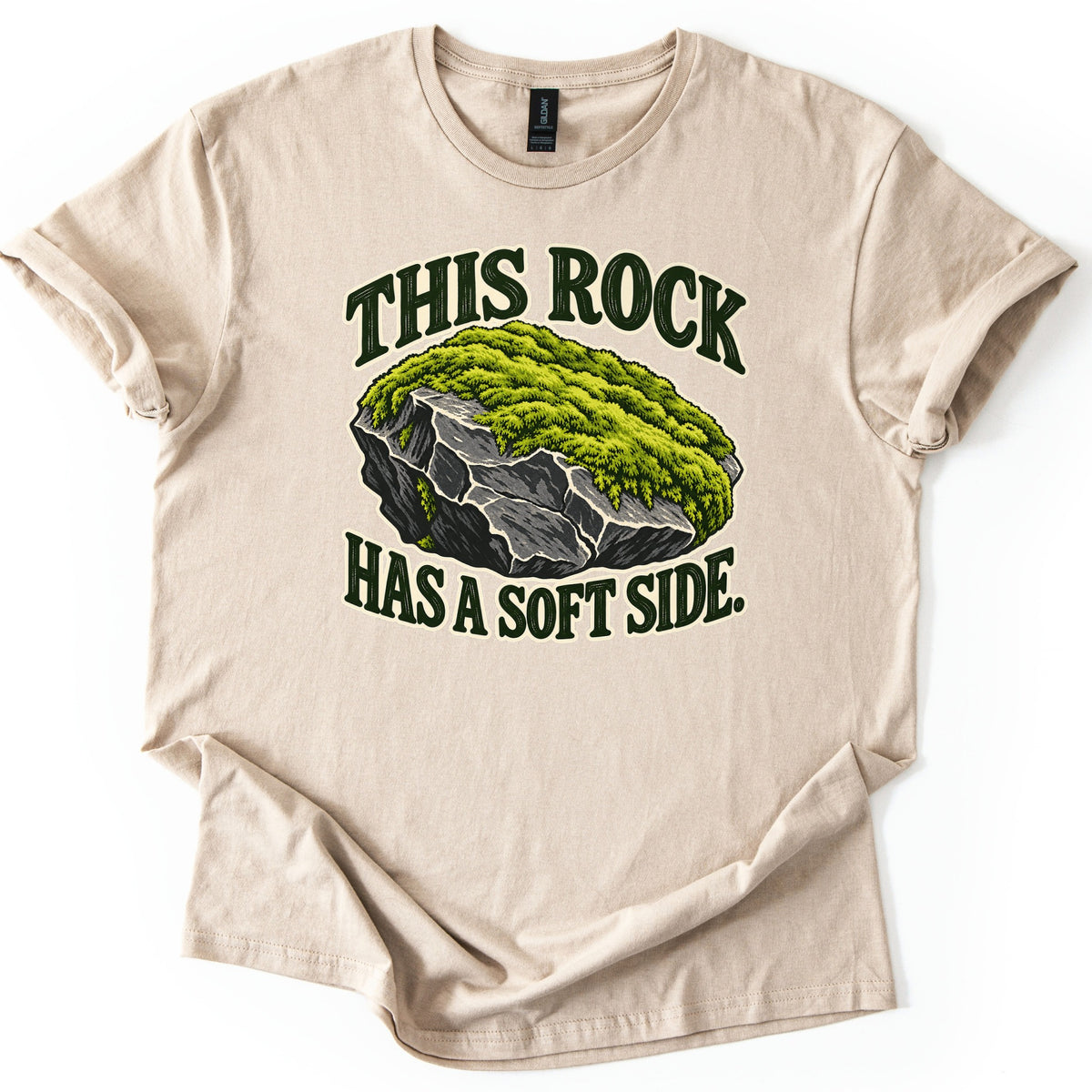 This Rock Has a Soft Side Comfort Colors T-shirt in Sand, laid flat. A gray boulder topped with thick green moss sits between the words This Rock above and Has a Soft Side. below in dark green letters with a cream outline. The shirt lies on white with cuffed sleeves.