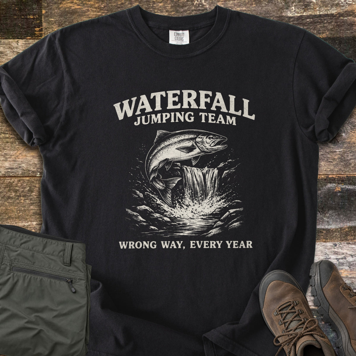 Waterfall Jumping Team Comfort Colors T-shirt in Black, laid flat. A salmon leaps beside a small waterfall between the words Waterfall Jumping Team and Wrong Way, Every Year, printed in cream. Shown on rustic wood with folded olive pants and brown hiking boots.
