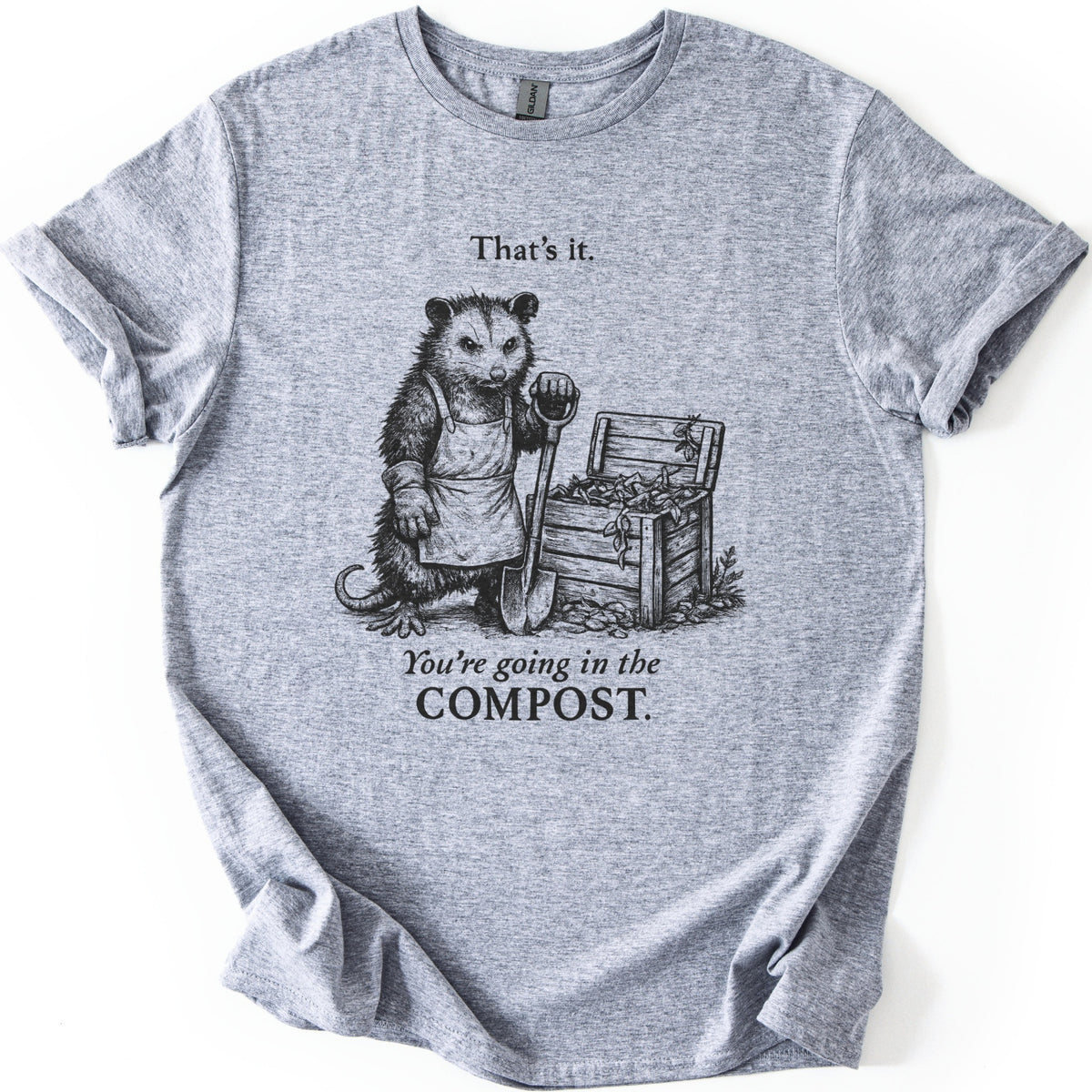 Going In The Compost Possum Comfort Colors T-shirt in Sport Grey, laid flat. A scowling possum in an apron and work glove leans on a shovel beside an open wooden compost bin, between the words That's it. and You're going in the COMPOST. Black print, shirt on white with cuffed sleeves.