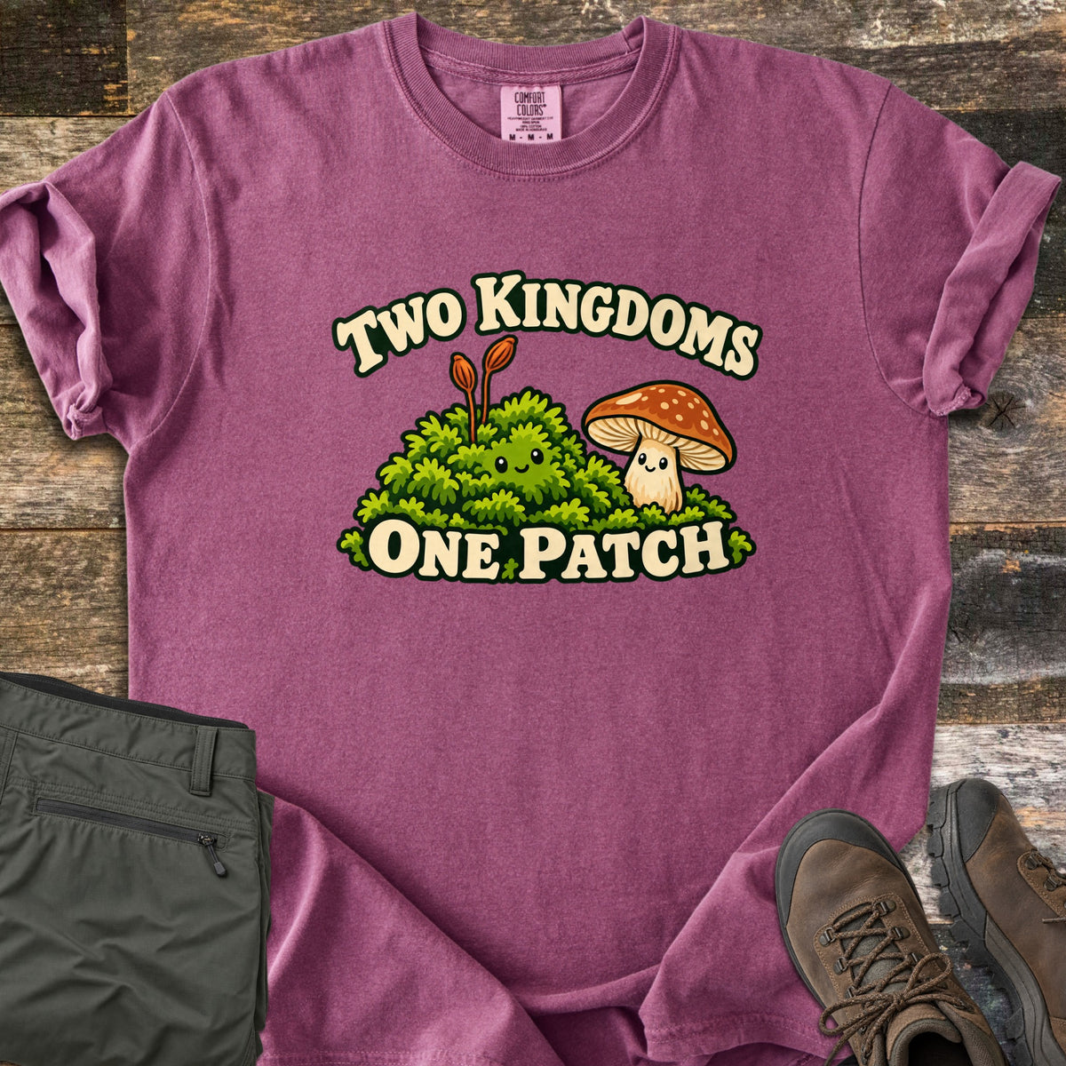 Two Kingdoms One Patch Comfort Colors T-shirt in Berry, laid flat. A smiling moss clump with two orange stalks sits beside a smiling spotted brown mushroom, between the words Two Kingdoms and One Patch. Sleeves rolled, on weathered wood with olive hiking pants and brown boots.