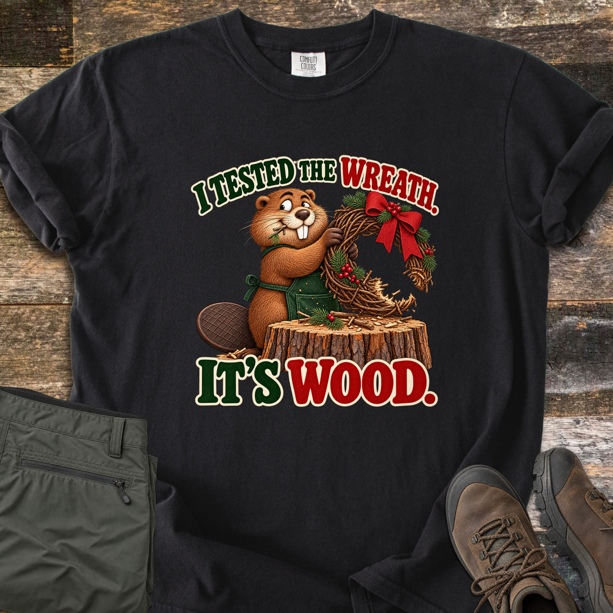 I Tested the Wreath. It's Wood. Comfort Colors T-shirt in Black, laid flat. A cartoon beaver in a green apron chews a twig wreath with a red bow on a tree stump, under the words I Tested the Wreath. It's Wood. Olive pants and brown hiking boots lie beside it on weathered wood planks.